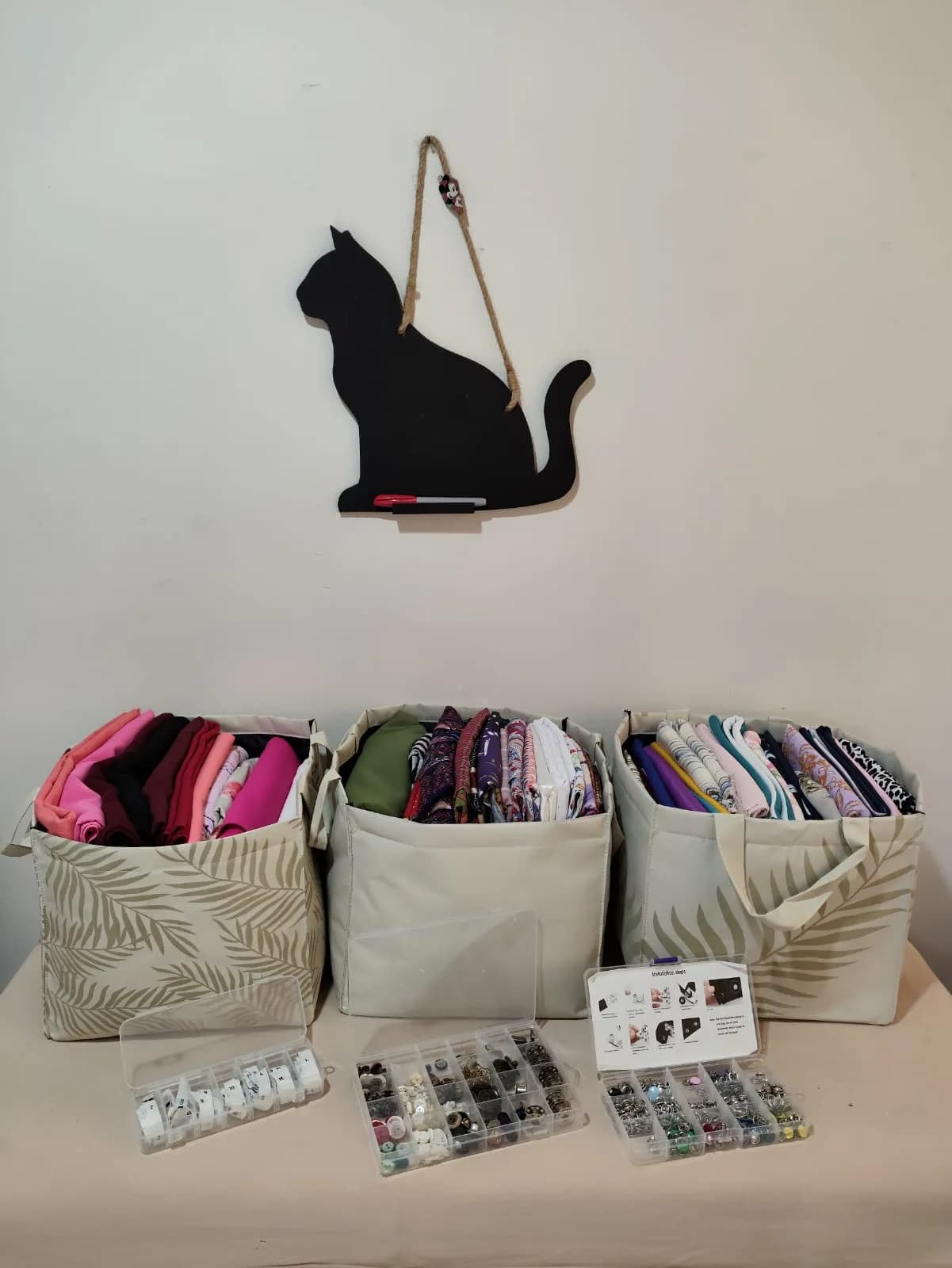 Fabric arranged in white baskets with clear containers of buttons and other sewing accessories with a black cat decoration hanging on wall